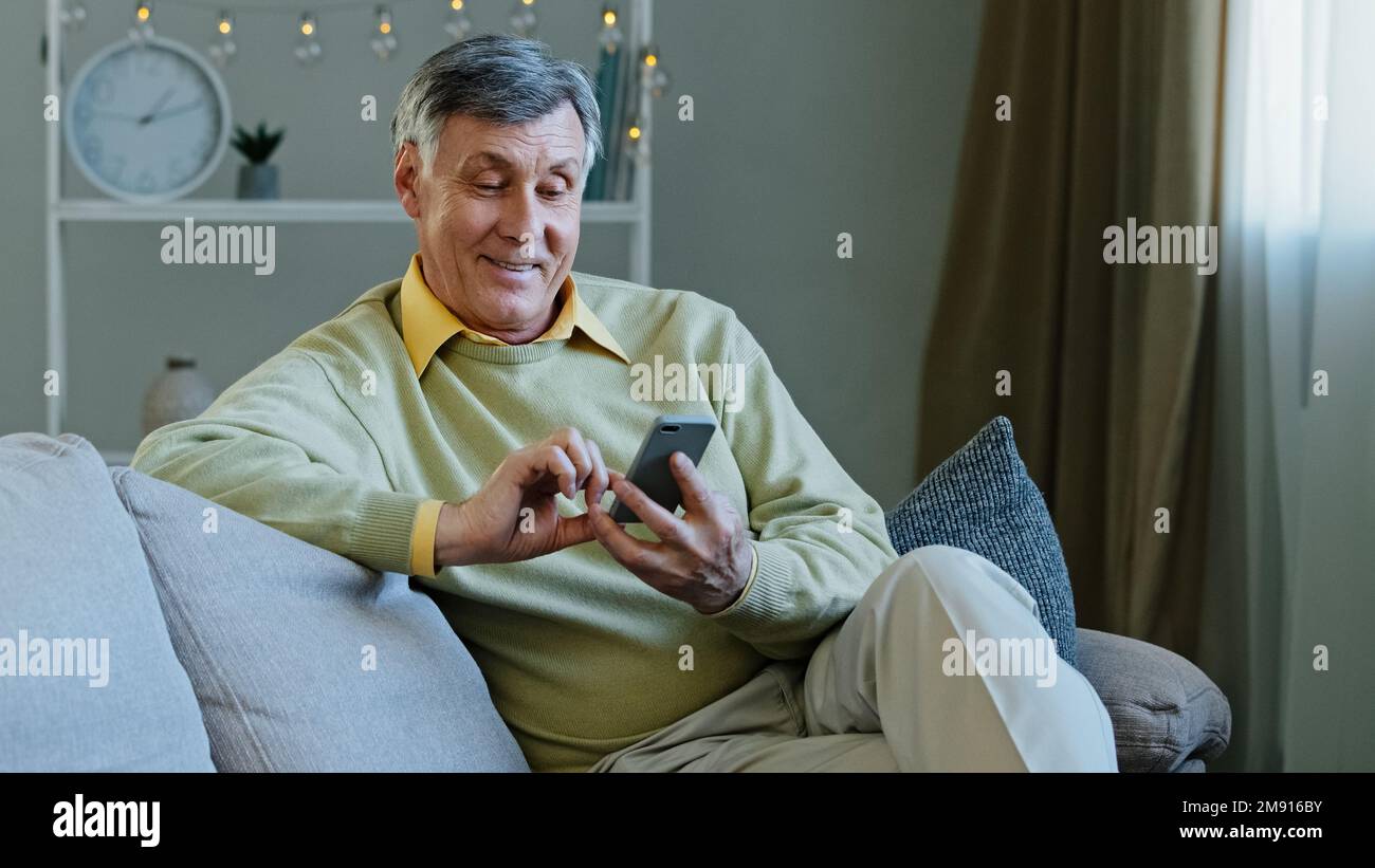 Caucasian elderly adult man sitting on sofa at home with gadget using online app on smartphone makes online order old grandfather resting reading news Stock Photo