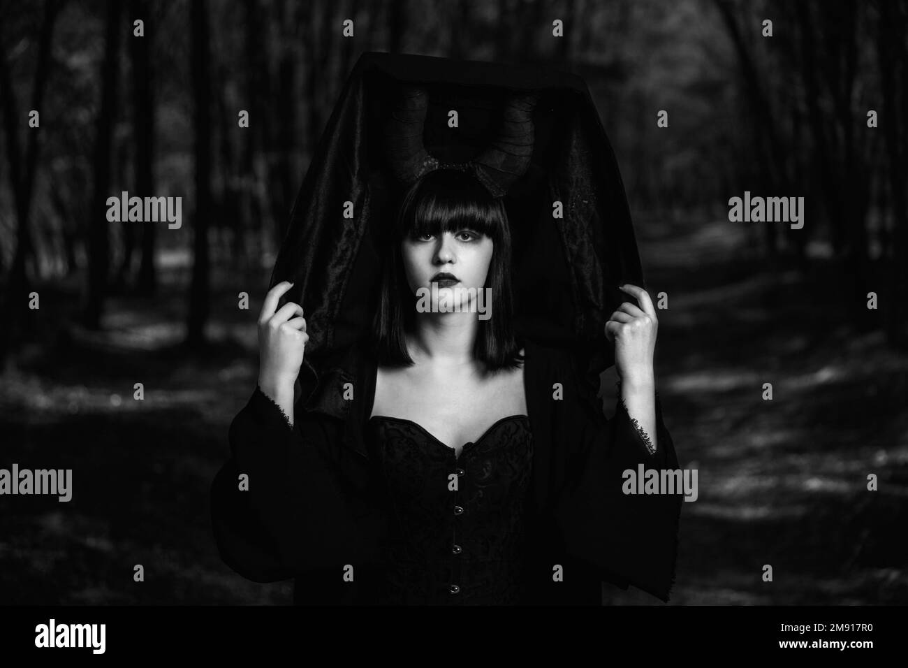 A grayscale of a gothic girl covering her Maleficent horns with a black cloak in a mystic forest Stock Photo