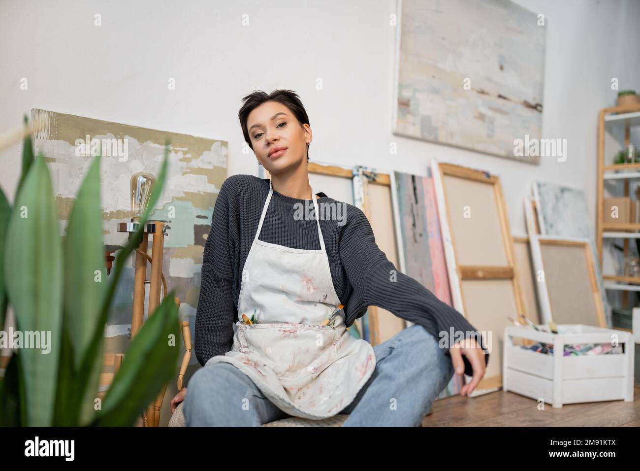 Young artist in apron looking at camera while sitting near plant and ...