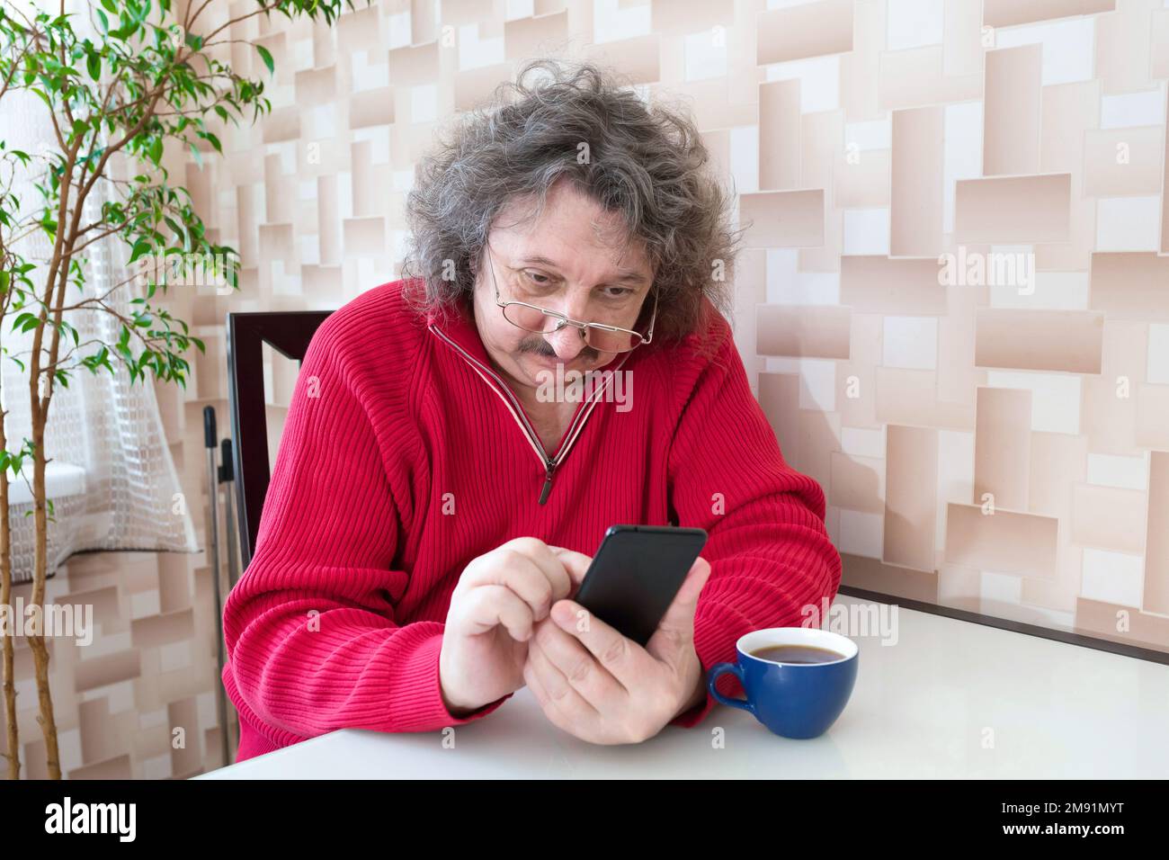 An elderly man in a red sweater and glasses with a phone in His Hands is sitting at the table. A pensioner, ageing Stock Photo