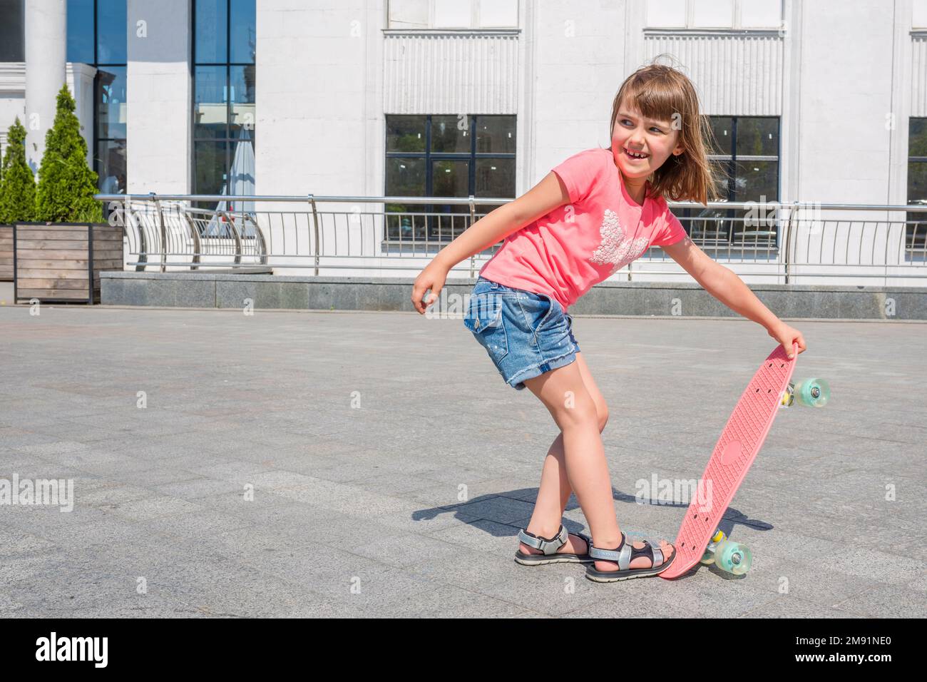 An outdoors portrait of a happy little girl with a skateboard on a summer sunny day in the park on a river bank Stock Photo