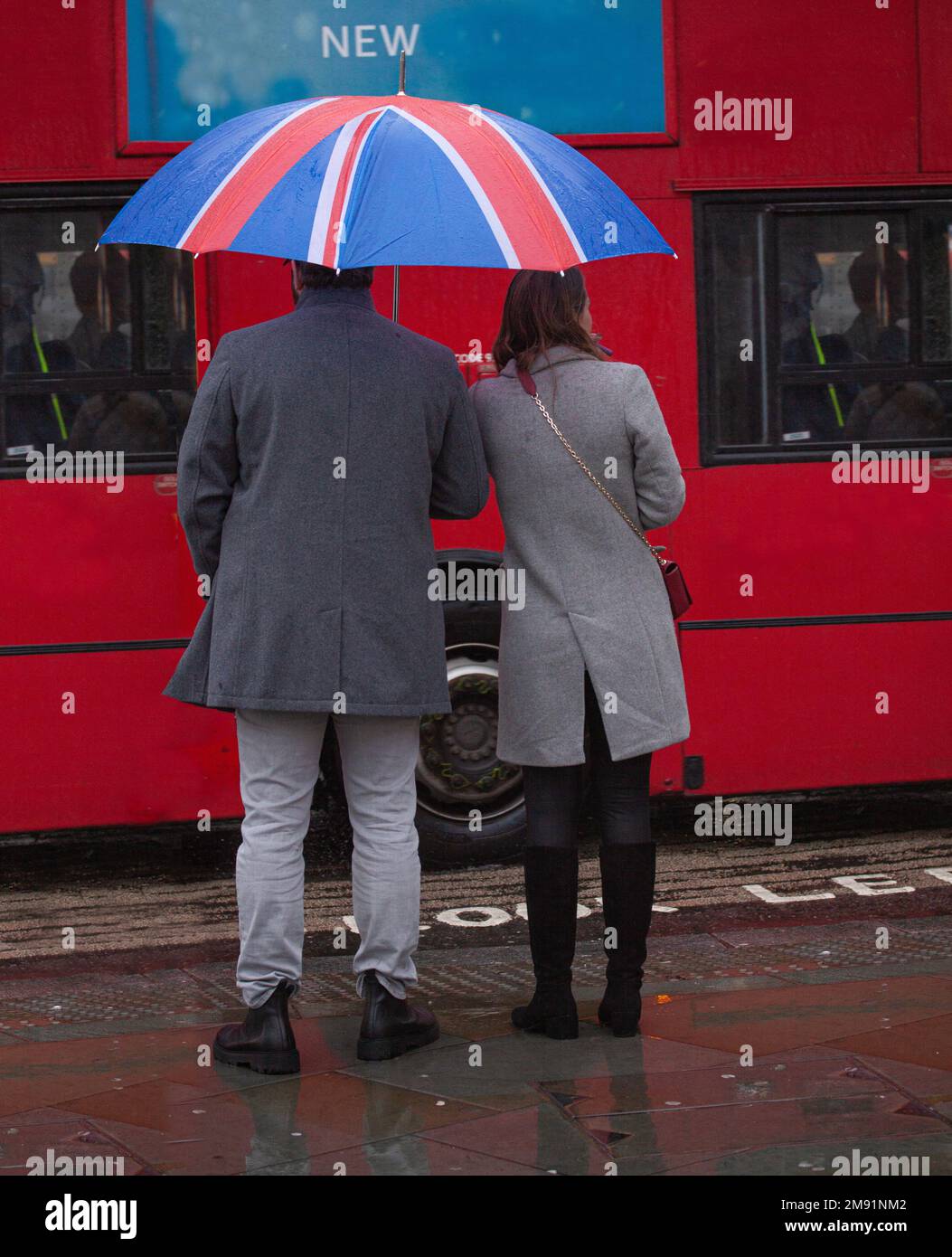 Back view of well-dressed young couple with British flag umbrella wainting in front af a red London double-decker bus. London, England - December 31, Stock Photo