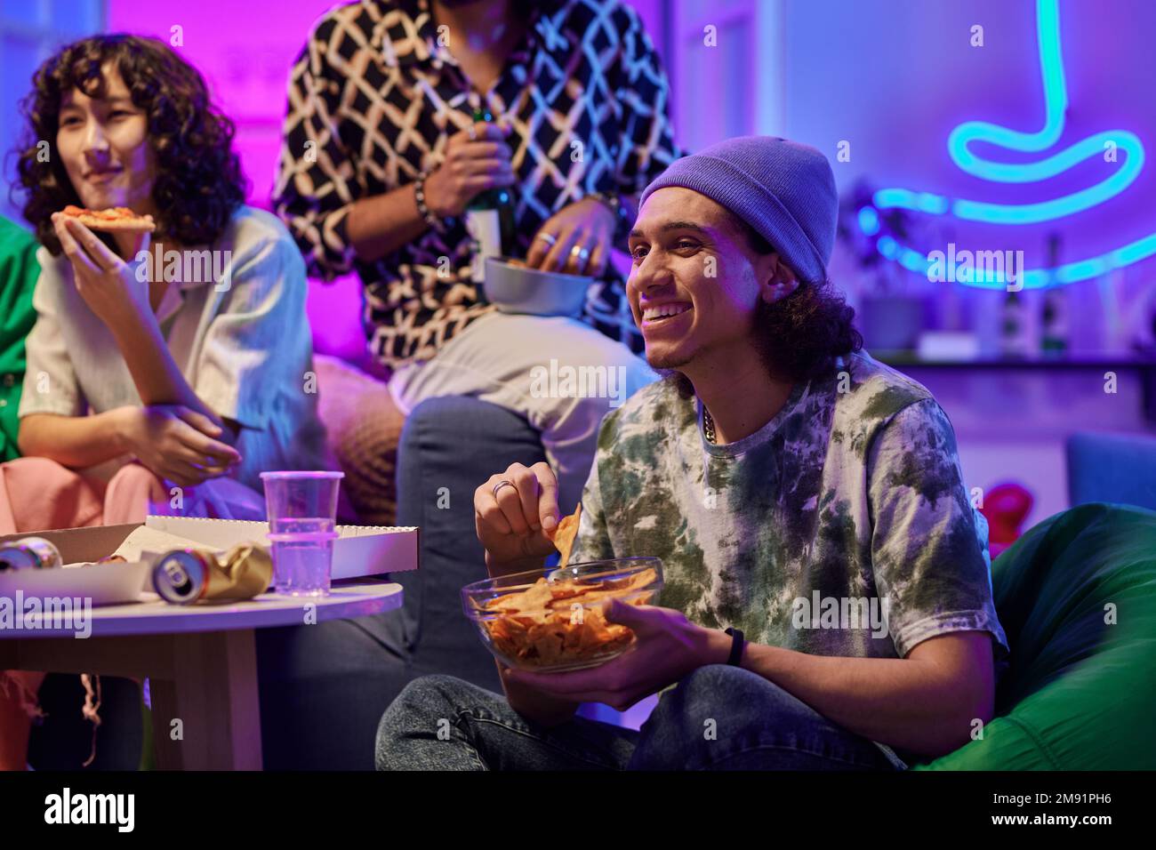 Happy young man eating potato chips and watching movie on tv while sitting against his friends in living room lit by neon light Stock Photo