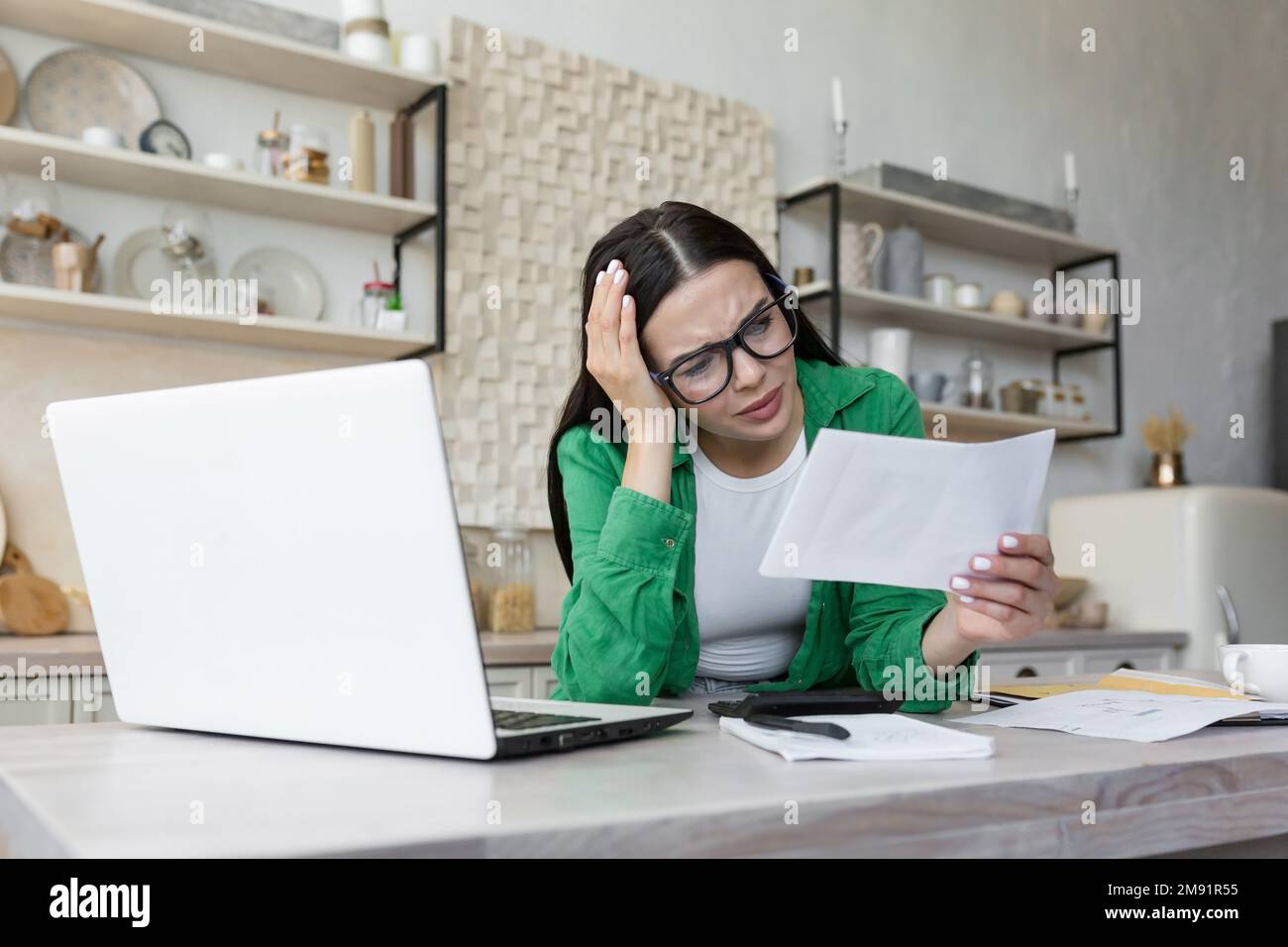 Shocked young woman working in kitchen at table with laptop. She holds a letter, a document in her hand, received bad news, an account, debt, divorce papers. He holds his head worriedly. Stock Photo