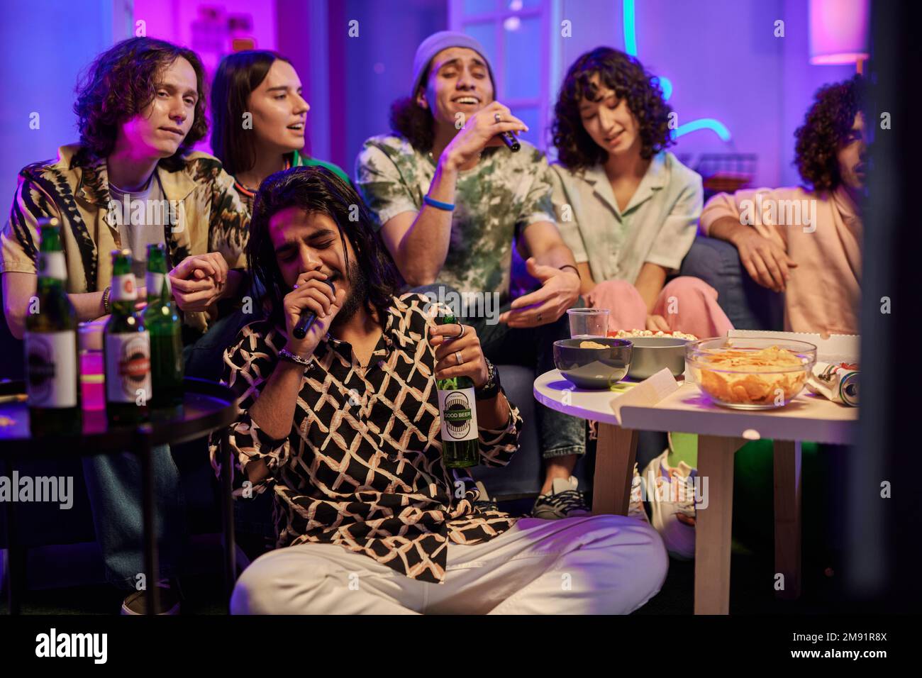 Two intercultural guys with microphones singing romantic song in karaoke while sitting among their friends listening to them at home party Stock Photo