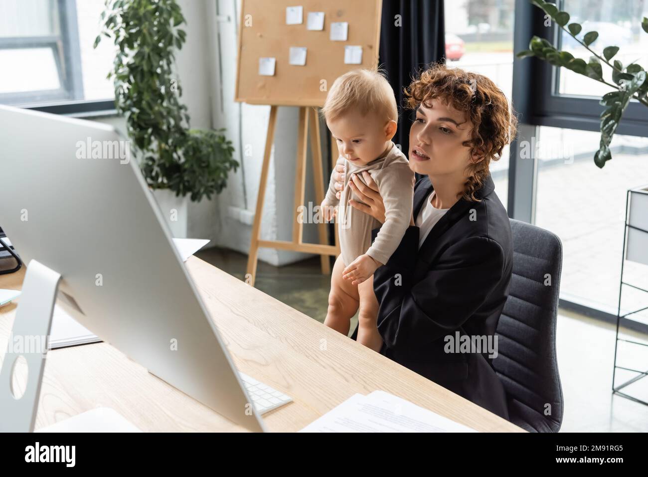 businesswoman in black blazer holding baby and looking at computer monitor in office,stock image Stock Photo