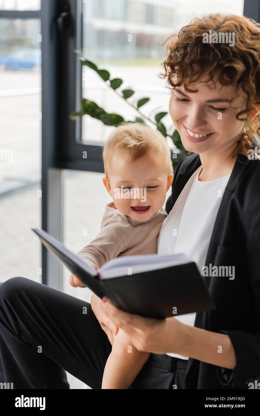smiling businesswoman holding cheerful toddler daughter and looking in notebook in office,stock image Stock Photo
