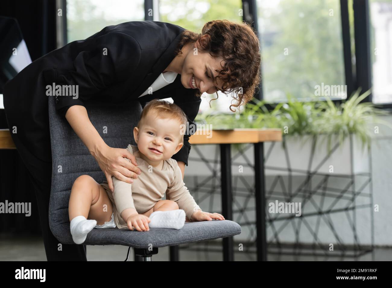cheerful baby in romper sitting on office chair near smiling mother in formal wear,stock image Stock Photo
