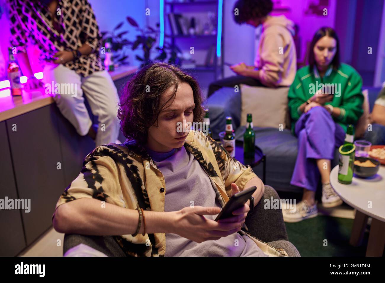 Serious guy in casualwear watching online video or scrolling through new posts in smartphone while relaxing among friends at home party Stock Photo