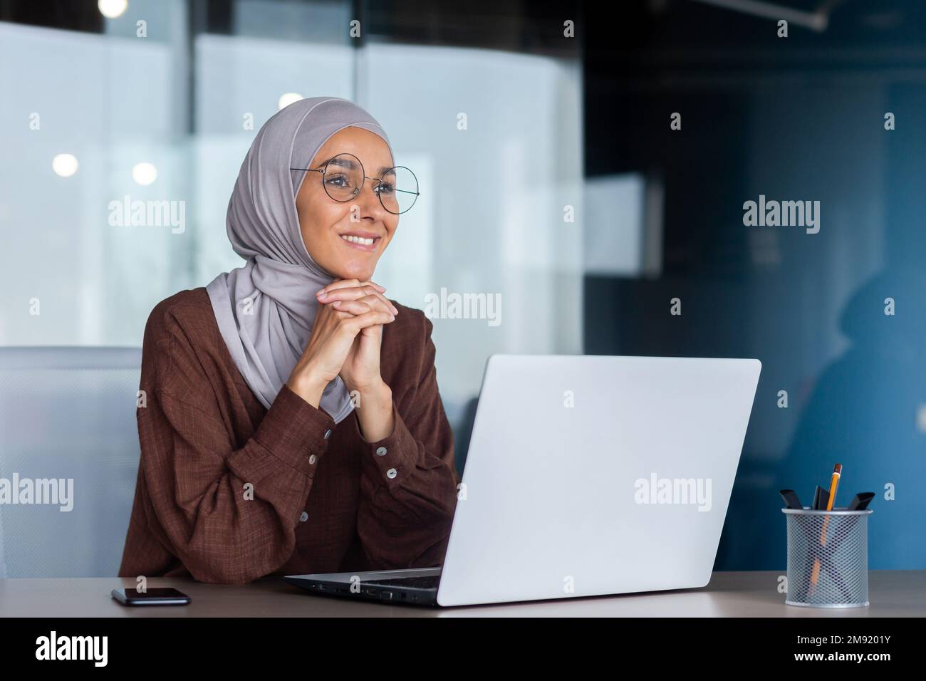 Smiling and dreamy businesswoman working inside office with laptop, woman in hijab and glasses office worker happy and satisfied with work sitting at desk. Stock Photo