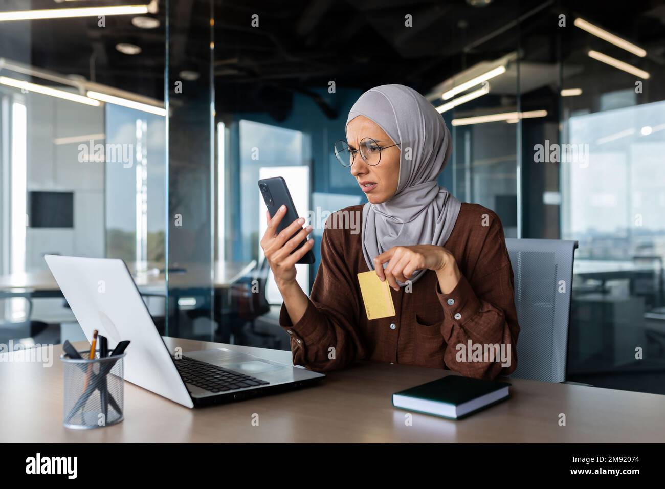 Upset and disappointed woman in hijab inside office trying to make purchase in online store, business woman holding smartphone and bank credit card, received money transfer error. Stock Photo