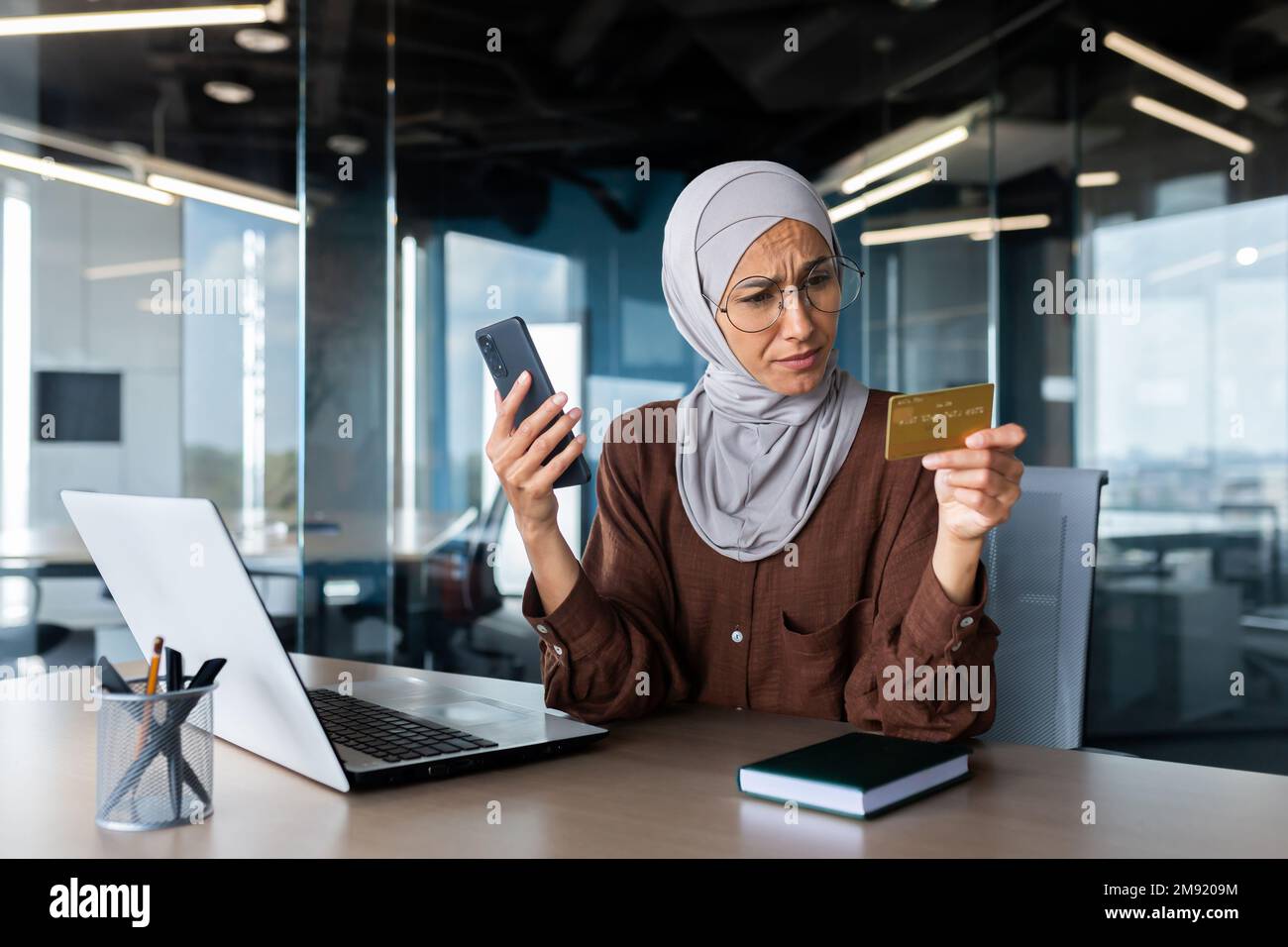 Upset and disappointed woman in hijab inside office trying to make purchase in online store, business woman holding smartphone and bank credit card, received money transfer error. Stock Photo