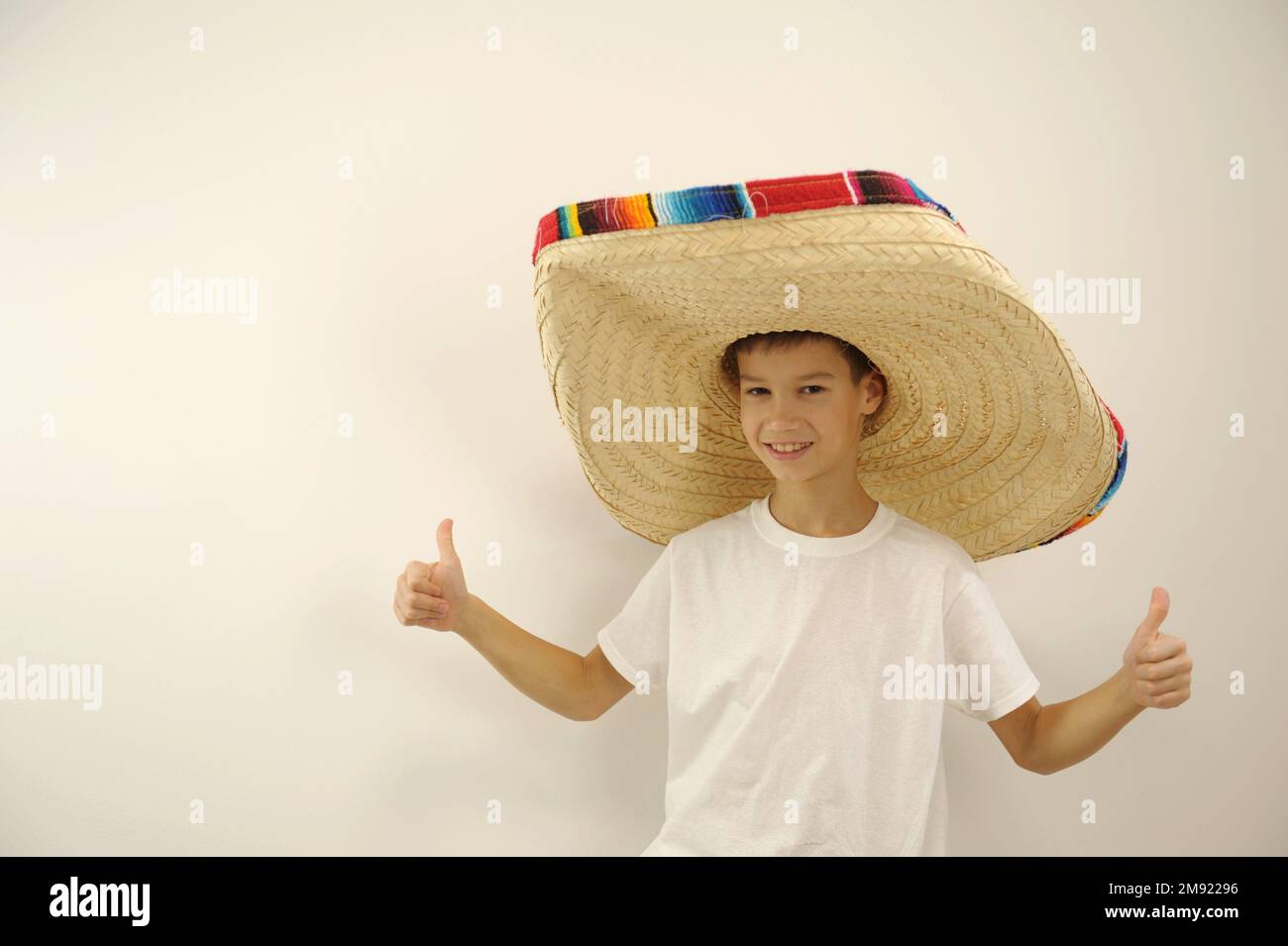 boy in a huge mexican hat shows thumbs up on both hands class he approves something place for text on a white background Young Mexican patriot with a ranger hat celebrating El Grito de Independencia Stock Photo