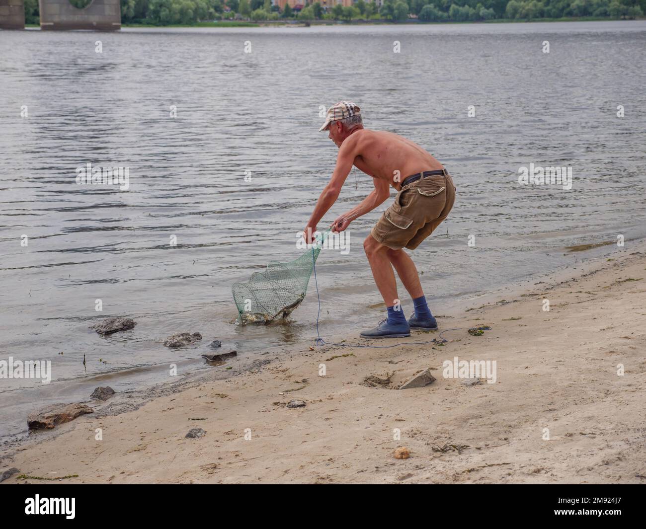 An old man in shorts and a cap holding a landing net in his hands and pulling fish out of Dnipro river on an urban beach near the North bridge in Kyiv Stock Photo