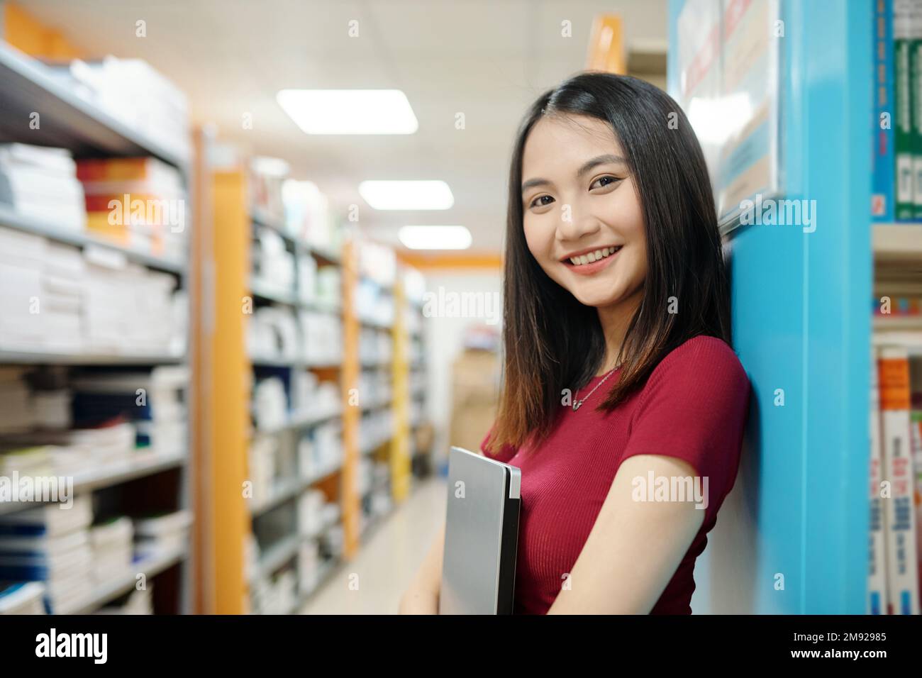 Portrait of smiling high school student with laptop standing in library ...