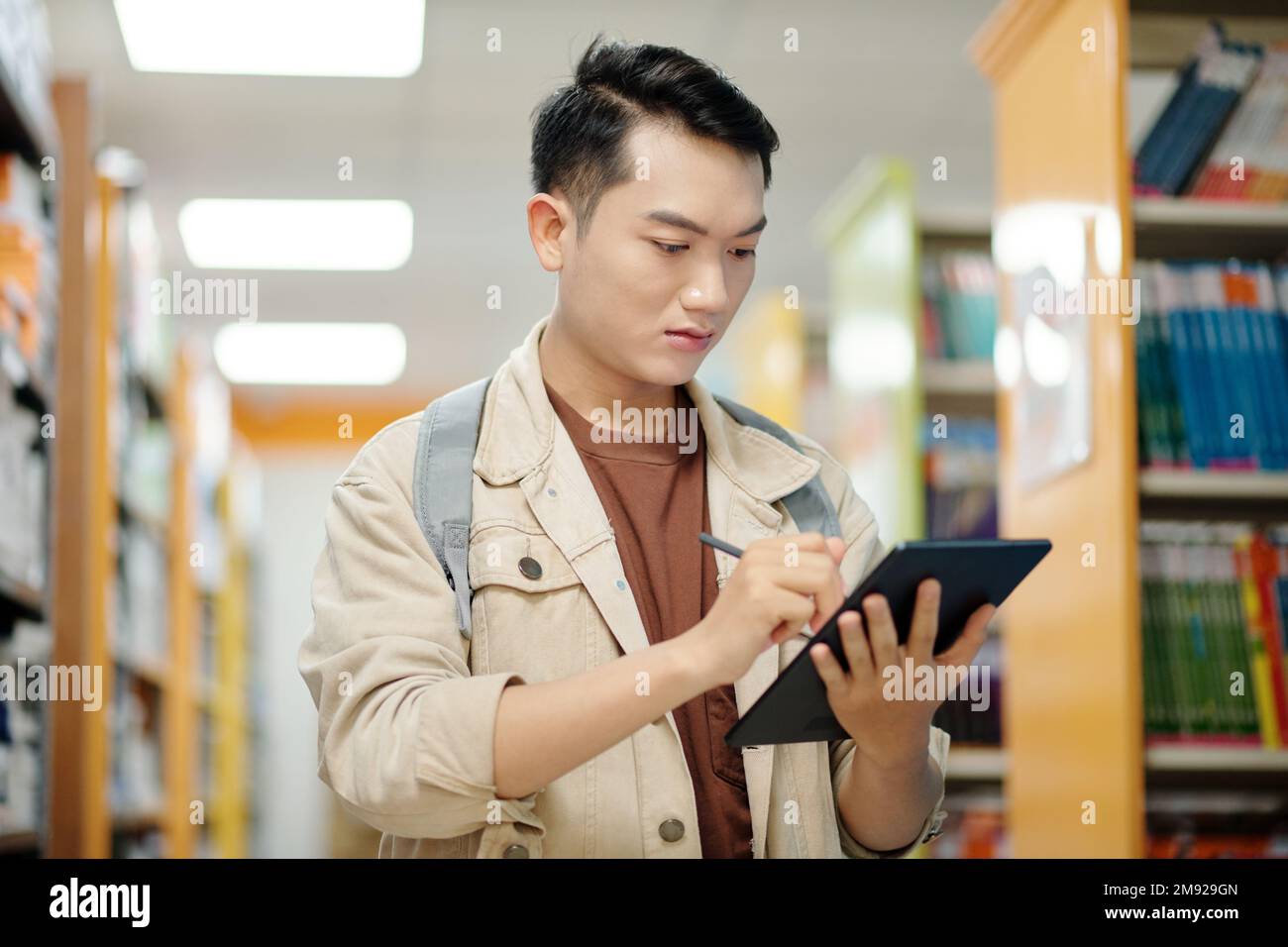 Serious college student standing in library and taking notes on tablet computer Stock Photo