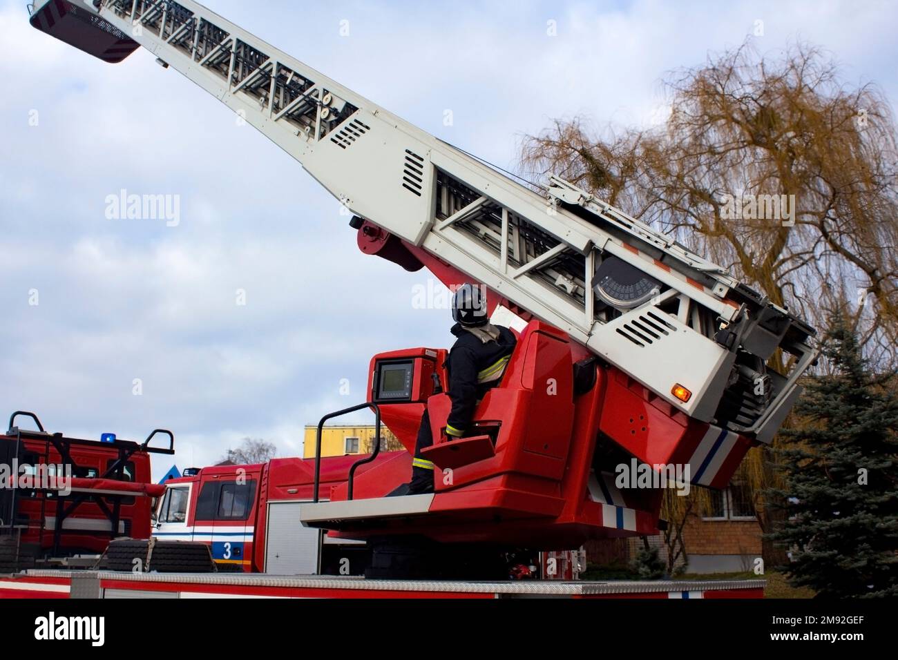 High retractable fire ladder with stall against blue sky, firefighter's rescue equipment. There is a rescuer in the control booth of the fire truck. Stock Photo