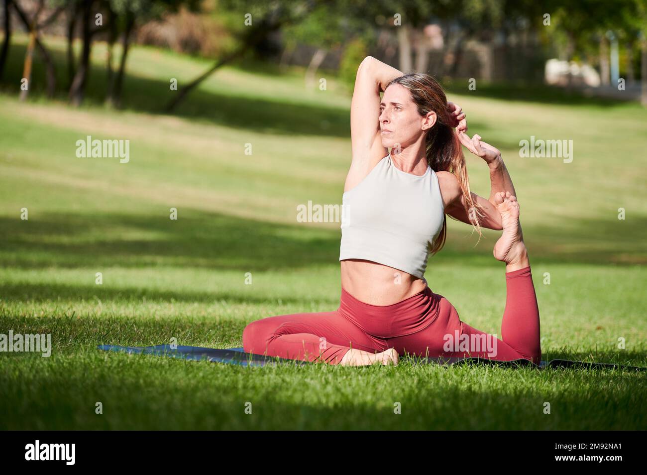 Full body of strong flexible sportswoman in activewear doing yoga in Eka Pada Raja Kapotasana during fitness training on green lawn Stock Photo