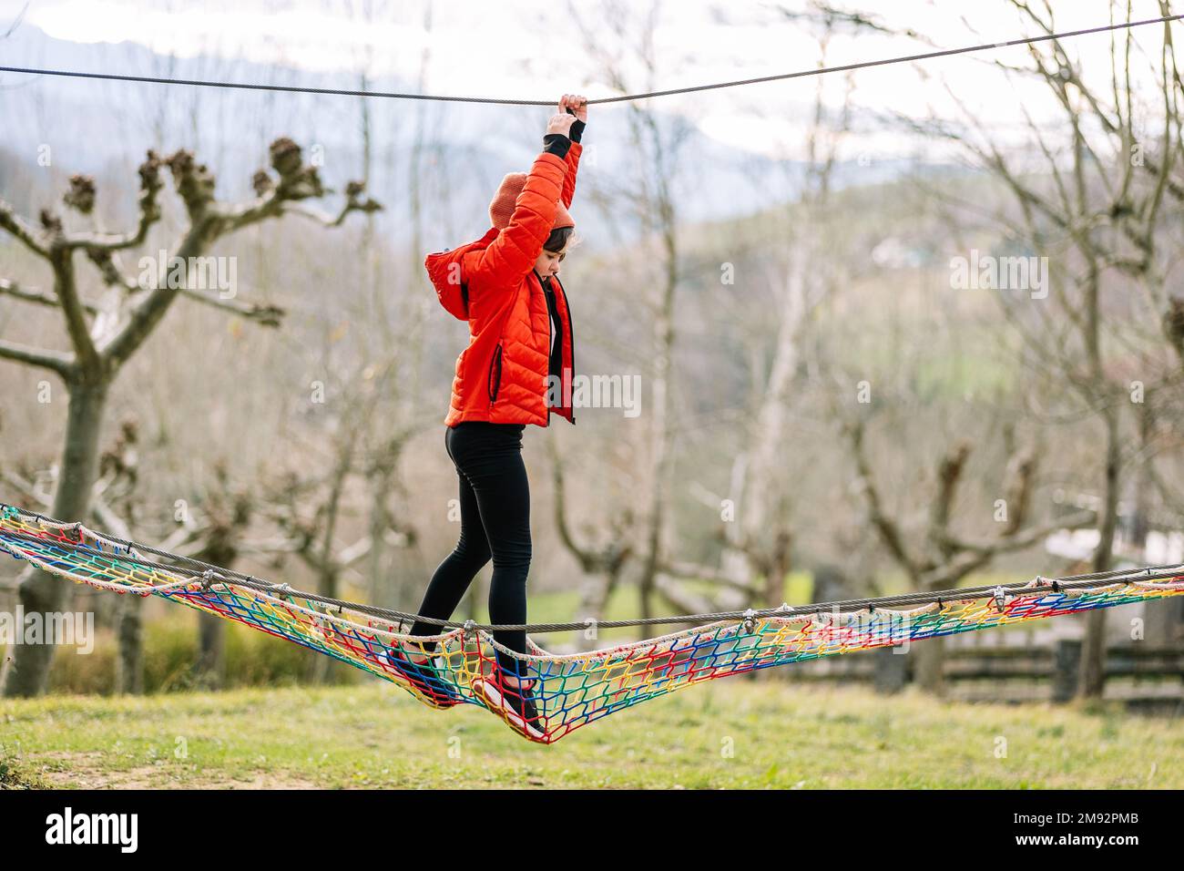 Side view full body of focused kid in warm clothes hanging on rope while walking on net in autumn park during weekend Stock Photo