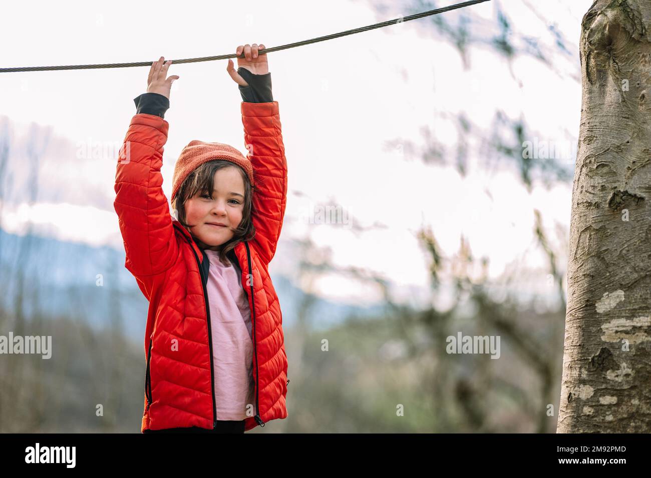Smiling kid in warm clothes hanging on rope while walking on net in autumn park during weekend Stock Photo