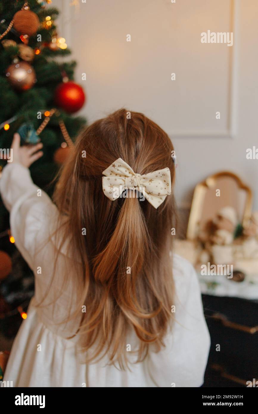 Back view of blond haired girl with bow standing against decorated Christmas tree while looking at Christmas bauble Stock Photo