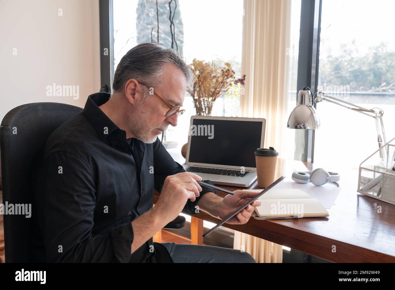 Side view of focused mature male entrepreneur in smart casual sitting in armchair while working on digital tablet at home office Stock Photo