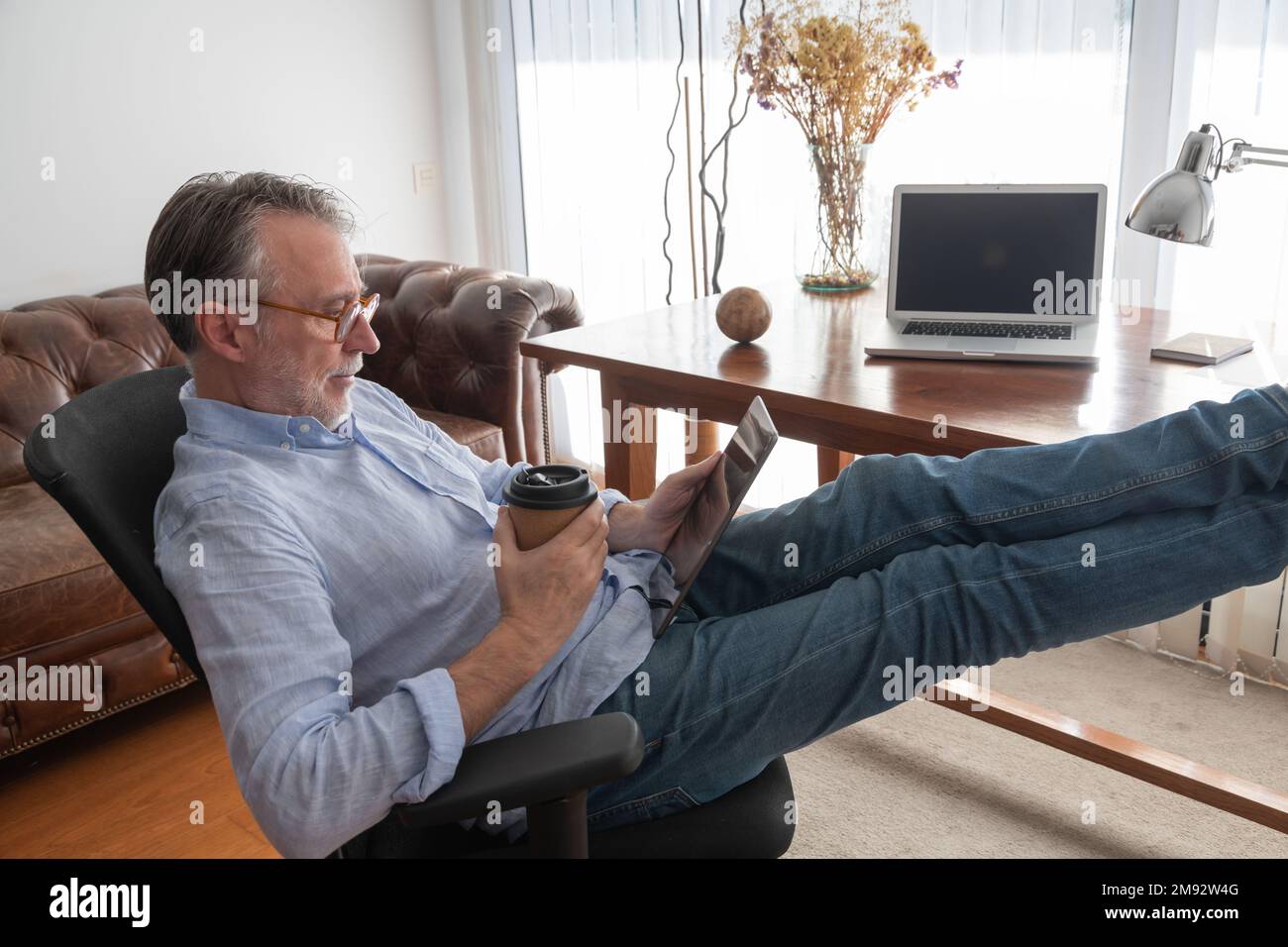 Side view of thoughtful man in casual clothes sitting in armchair with legs on table and drinking coffee while using digital tablet at workplace Stock Photo