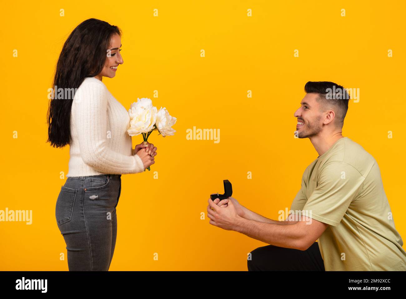 Glad millennial arabic male on knees give box with ring to smiling lady with bouquet of flowers Stock Photo