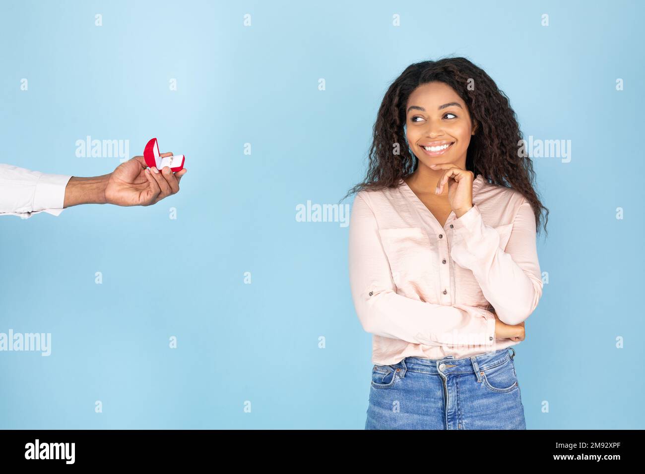 Happy african american woman looking at gift box with ring in man hand and smiling, standing on blue studio background Stock Photo
