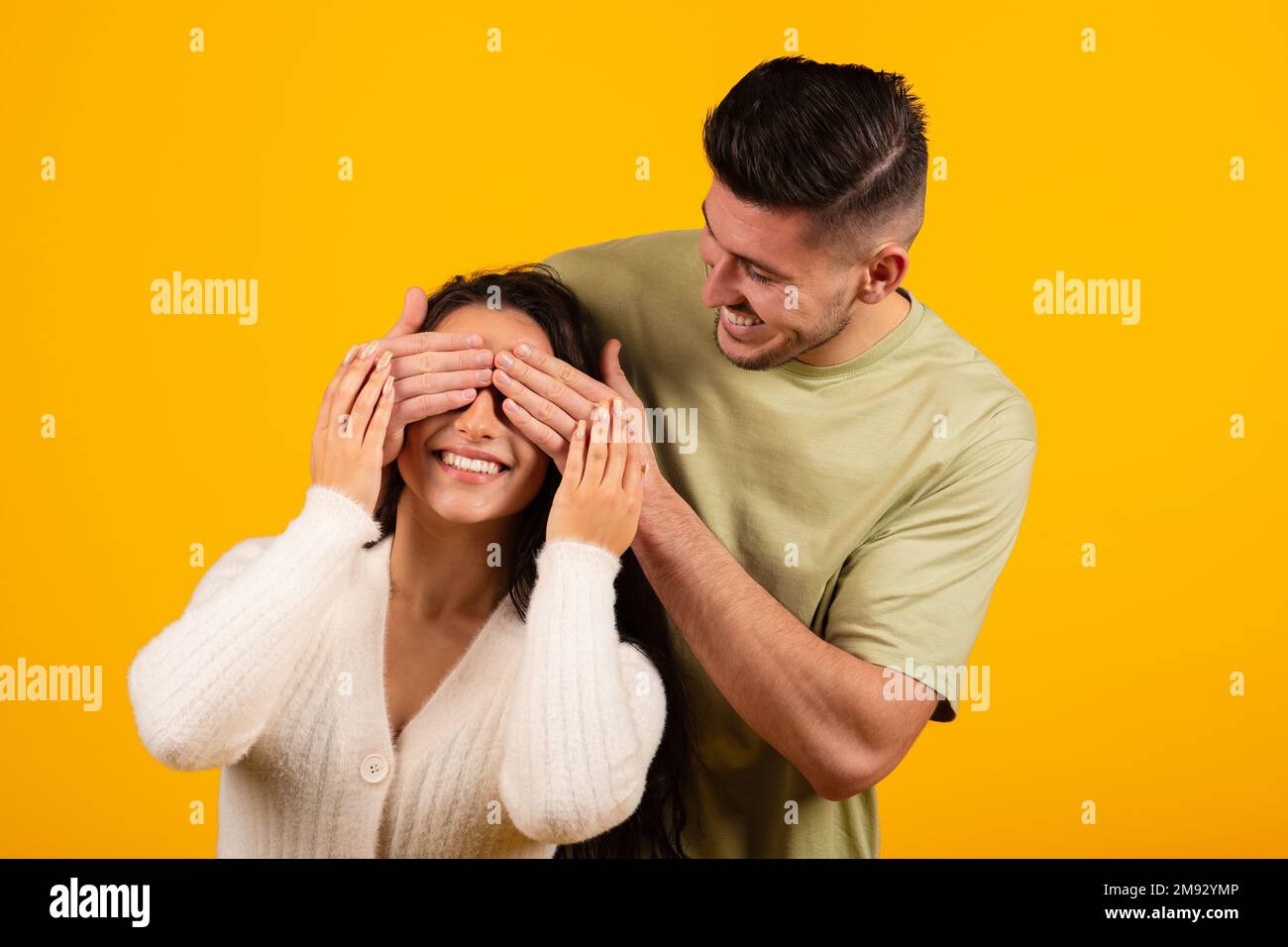 Smiling millennial arabic guy closes eyes of surprised lady, birthday greeting, isolated on orange background Stock Photo