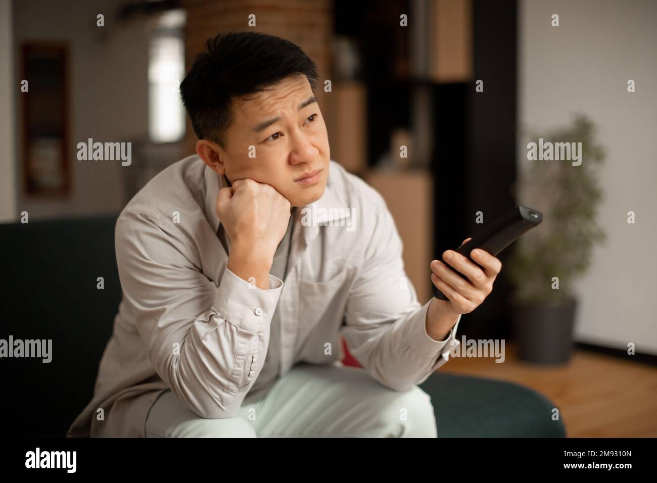 Boring television program concept. Bored mature japanese man watching TV, switching channels with remote control Stock Photo