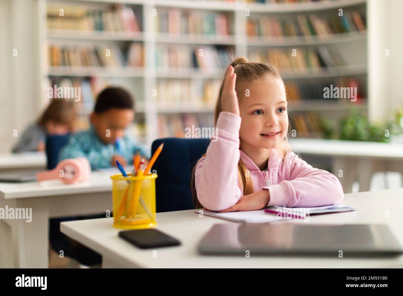 Education, elementary school concept. Pretty schoolgirl raising hand for an answer, sitting in classroom Stock Photo