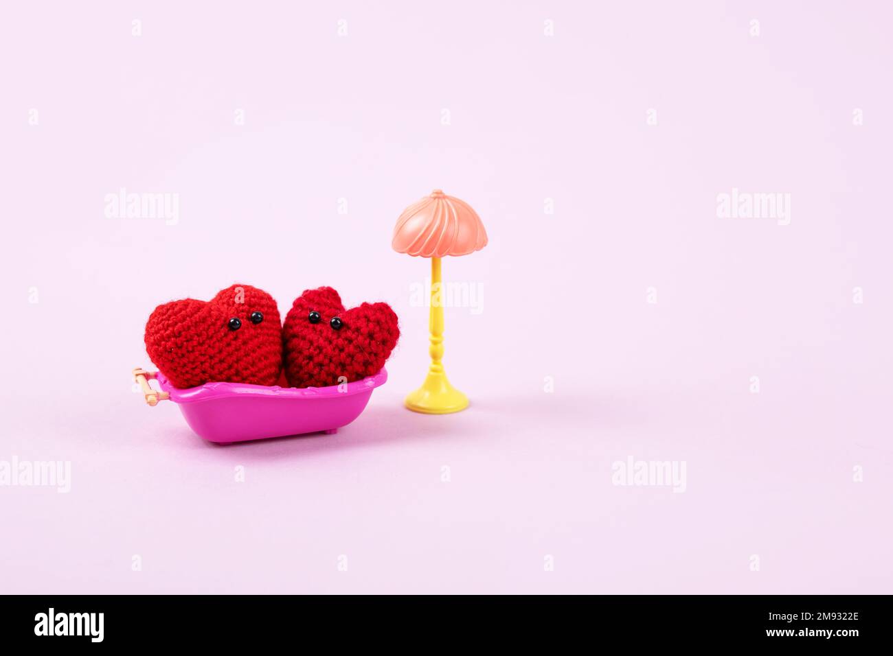 Concept of Valentine's Day. A family of two knitted red hearts in a pink toy bath and a toy lamp on a lilac background. Symbol of love, family, loyalt Stock Photo