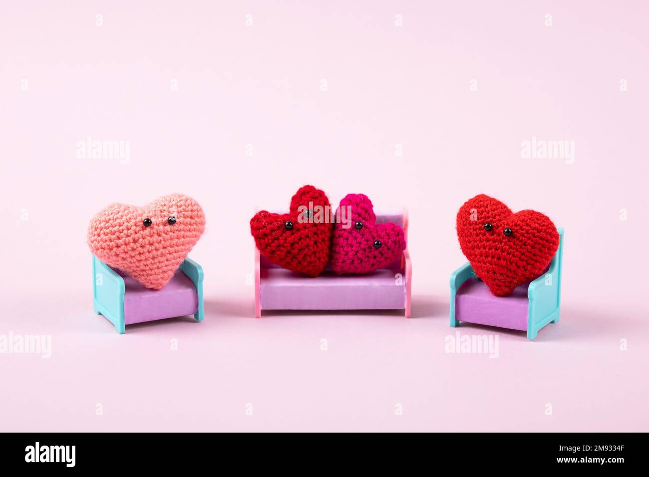 Concept of Valentine's Day. A composition from a family of knitted red and pink hearts that sit on toy chairs and a sofa on a lilac background. Symbol Stock Photo