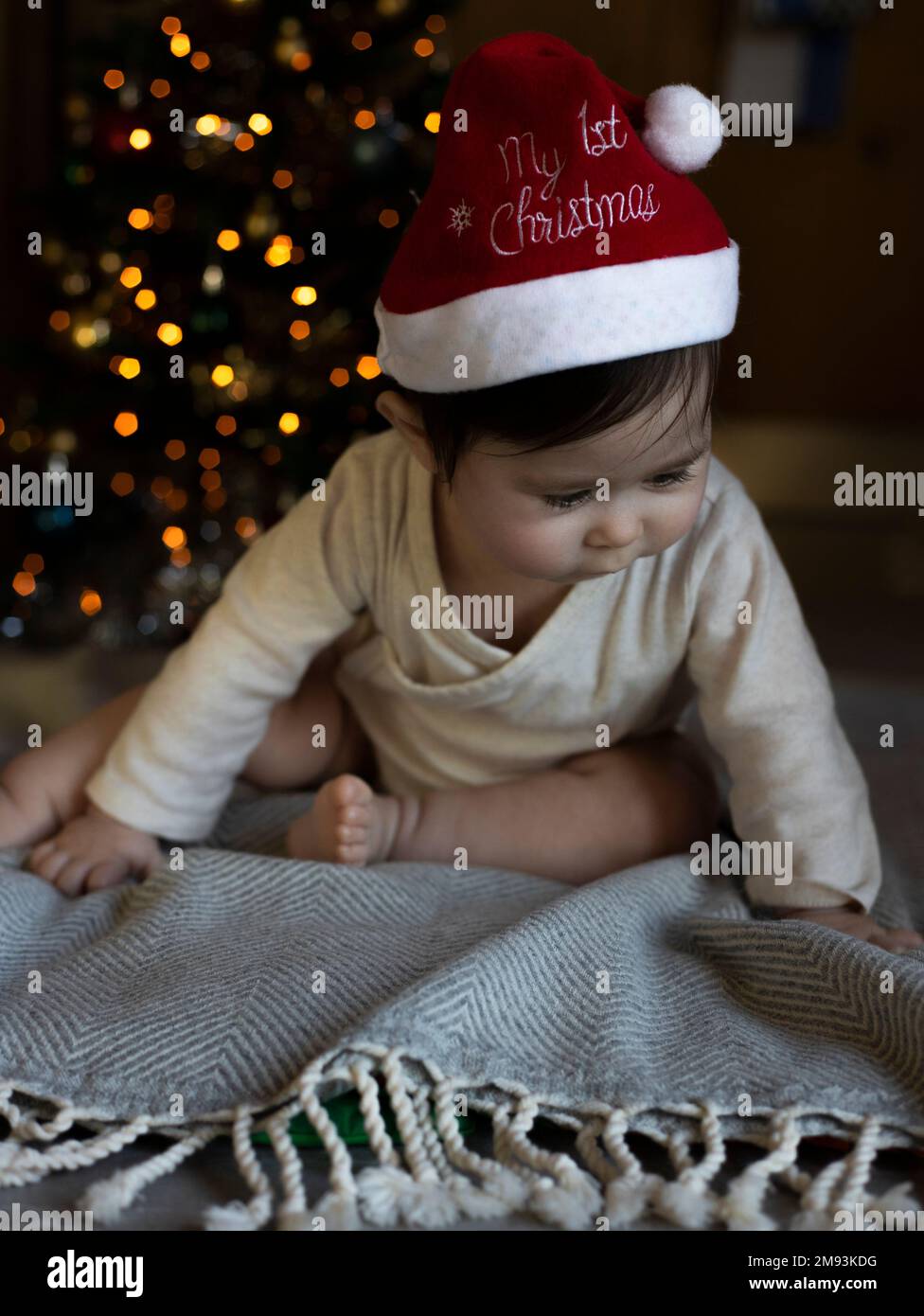 Adorable little baby wearing red christmas hat, sitting on soft blanket in front of blurry lights of christmas tree. Christmas celebration concept Stock Photo