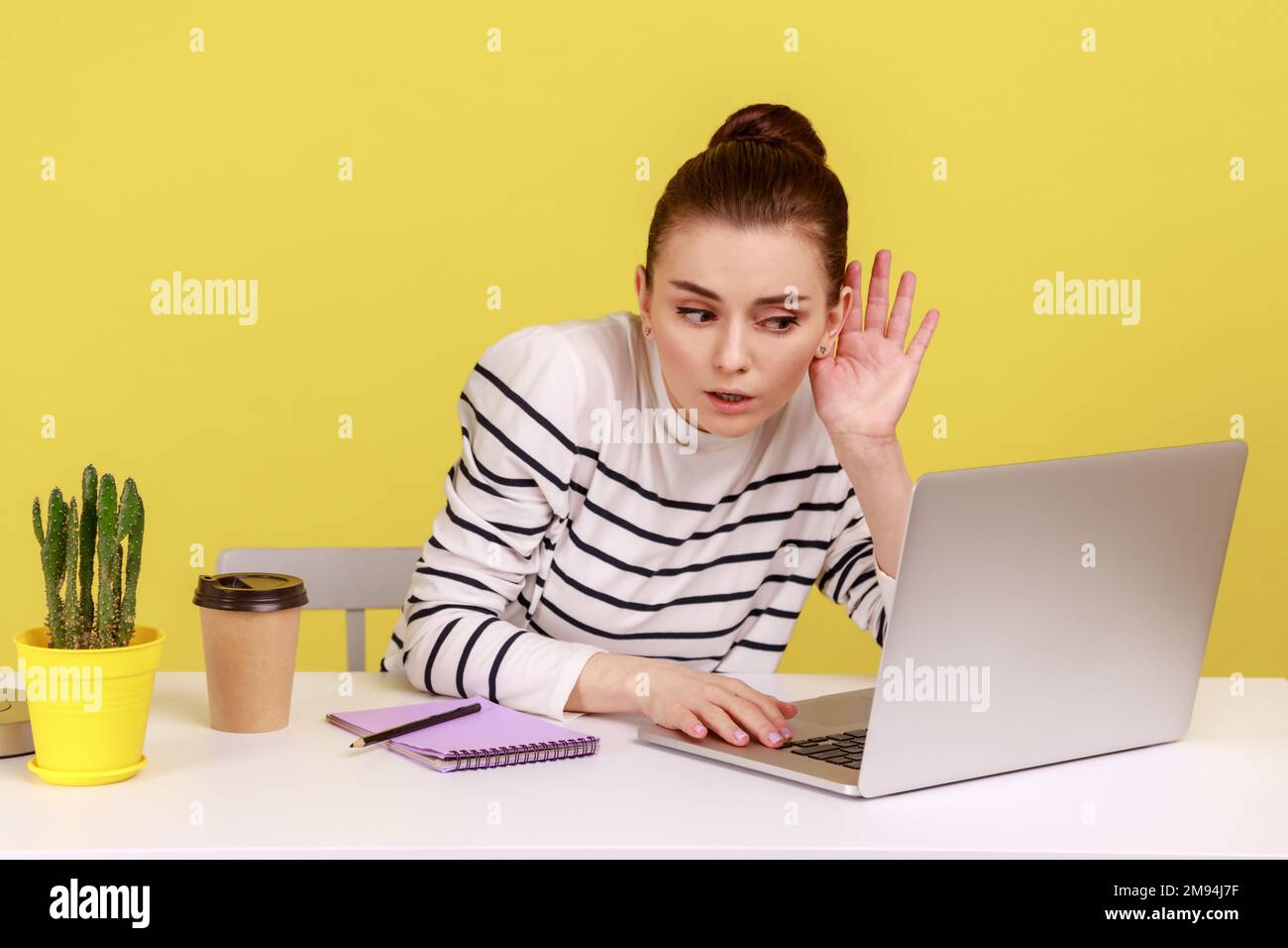 I can't hear you. Woman holding arm near ear trying to listen secret talk on video call on laptop, bad internet connection, online conference. Indoor studio studio shot isolated on yellow background. Stock Photo
