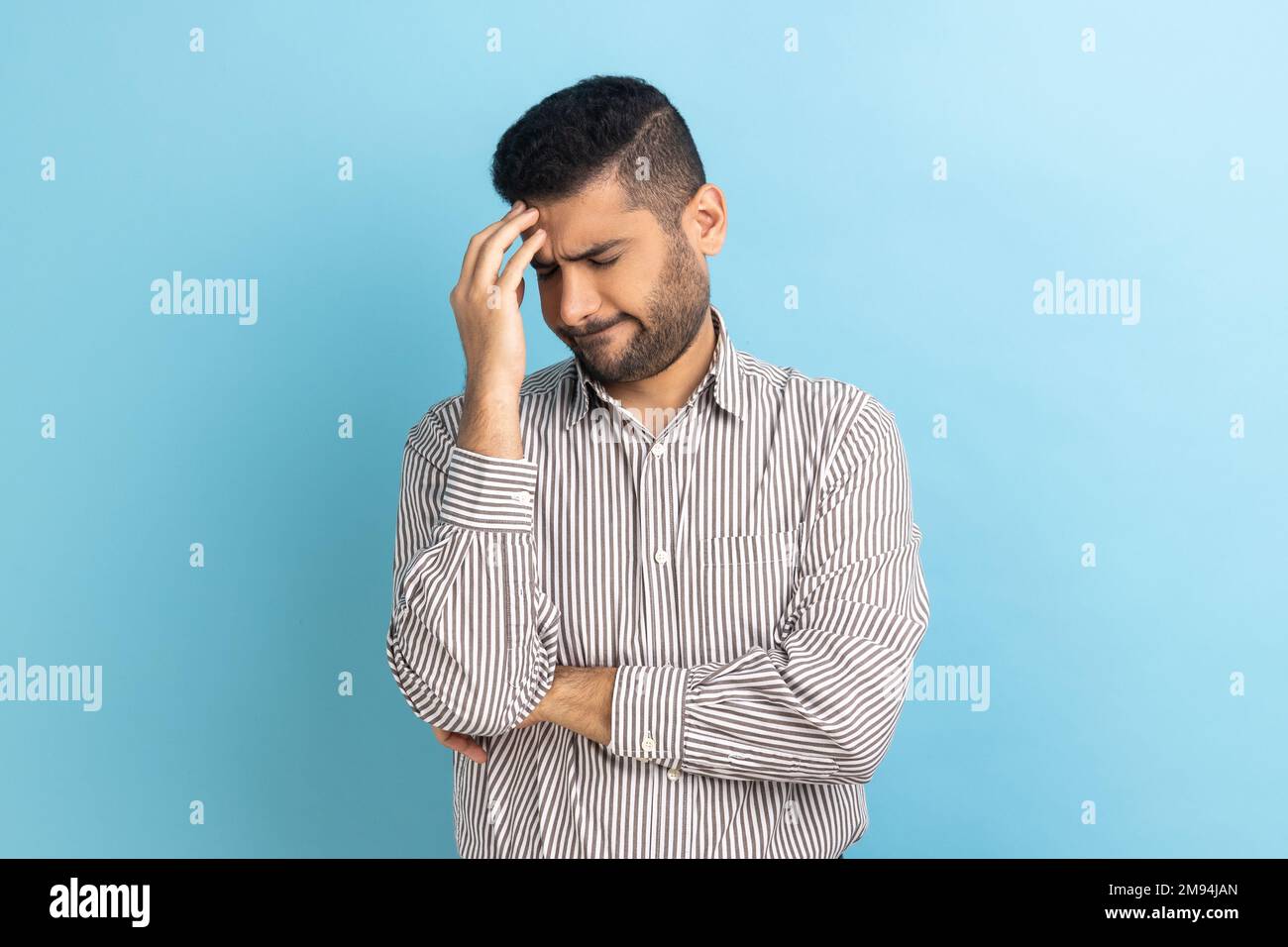 Businessman with beard making facepalm gesture keeping hand on head, blaming himself for bad memory, unforgivable mistake, wearing striped shirt. Indoor studio shot isolated on blue background. Stock Photo
