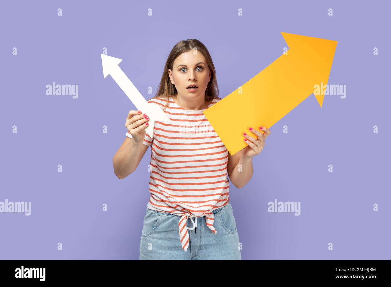 Portrait of shocked scared blond woman wearing striped T-shirt holding two arrows indicating to different sizes, looking at camera with big eyes. Indoor studio shot isolated on purple background. Stock Photo
