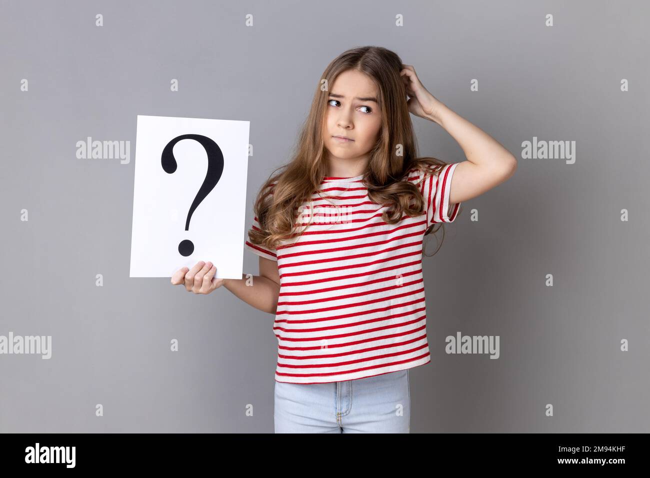 Portrait of little girl wearing striped T-shirt holding paper with question mark over, thoughtful, face thinking about question, very confused idea. Indoor studio shot isolated on gray background. Stock Photo