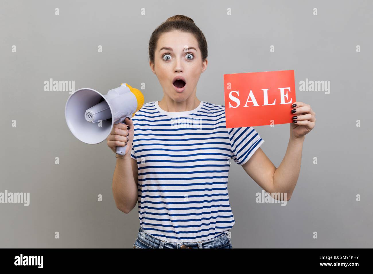 Portrait of shocked astonished amazed woman wearing striped T-shirt holding card with sale inscription and megaphone, making announcement. Indoor studio shot isolated on gray background. Stock Photo