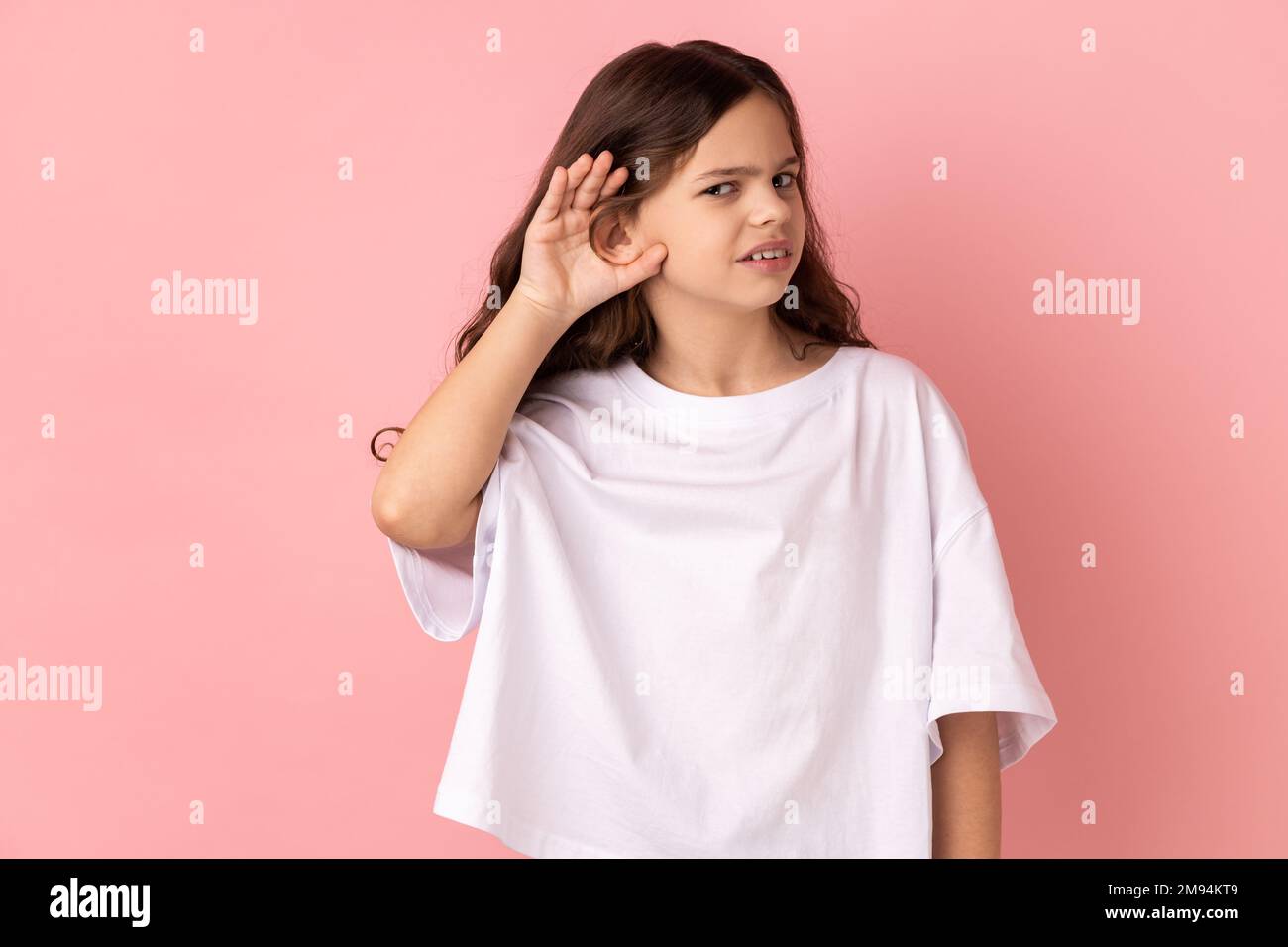 Portrait of curious little girl wearing white T-shirt holding hand near ear trying to hear secrets and find out gossips, listen to private whispers. Indoor studio shot isolated on pink background. Stock Photo