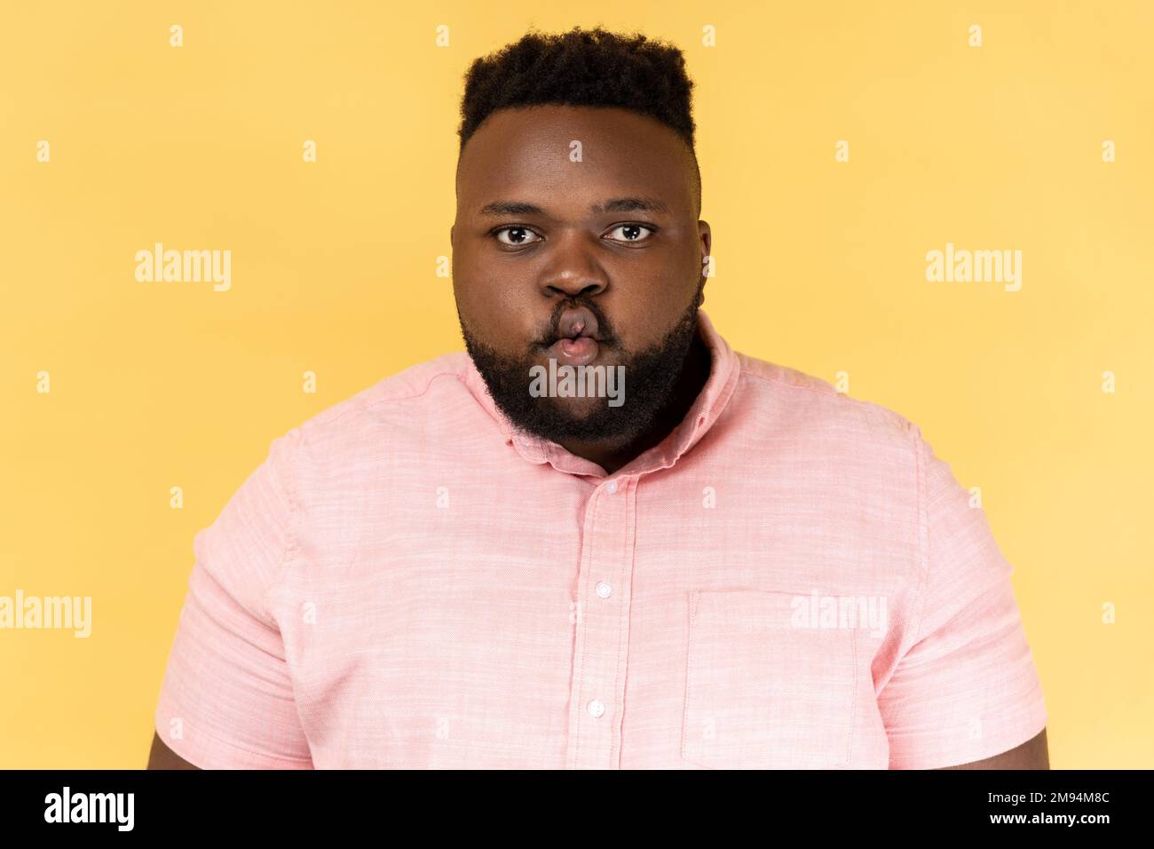 Portrait of funny positive young adult man wearing pink shirt standing with pouted lips making fish face, having fun, childish good mood. Indoor studio shot isolated on yellow background. Stock Photo
