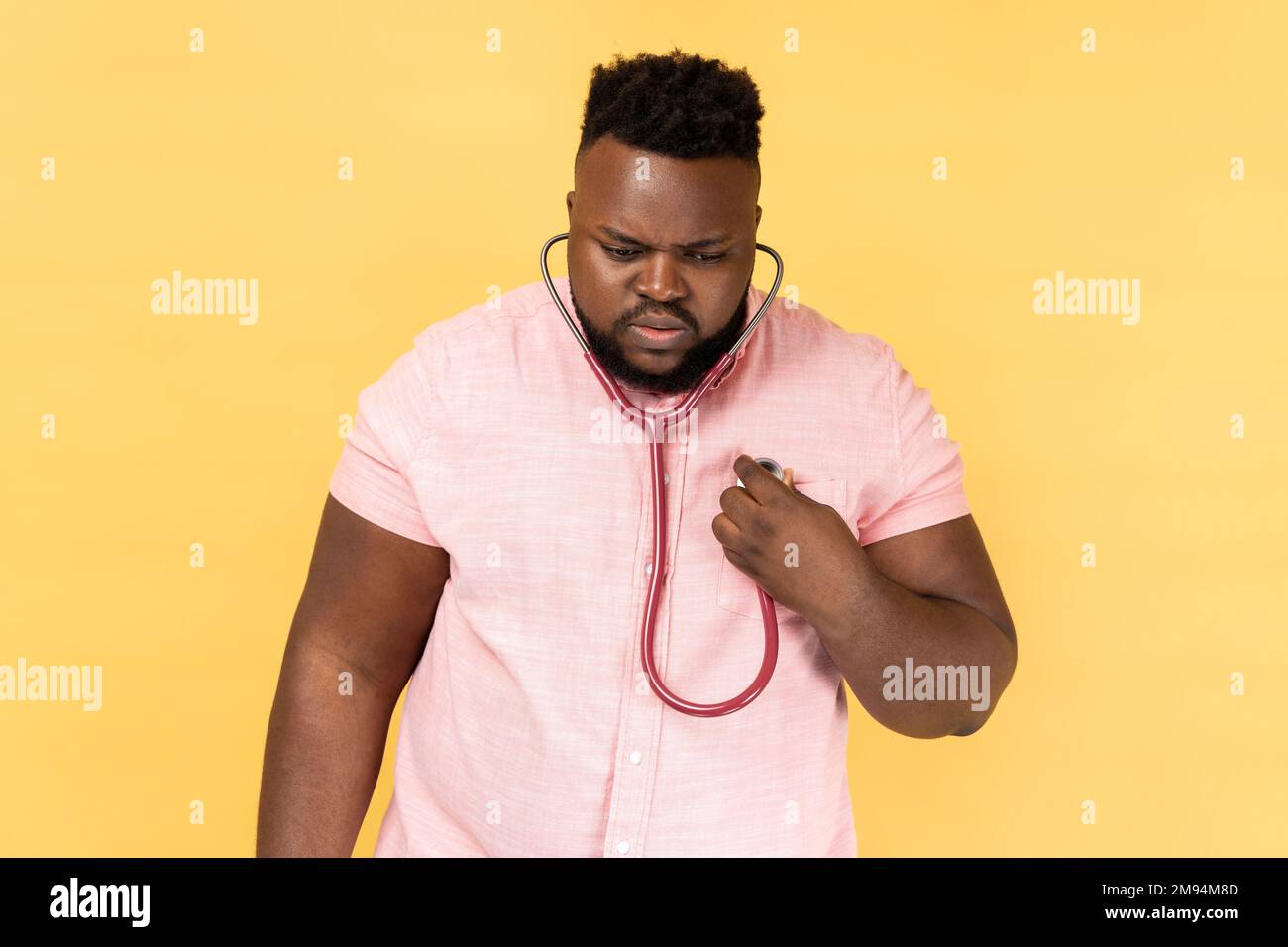 Portrait of concentrated bearded man wearing pink shirt standing with stethoscope, doing self examining of his heart, medicine concept. Indoor studio shot isolated on yellow background. Stock Photo