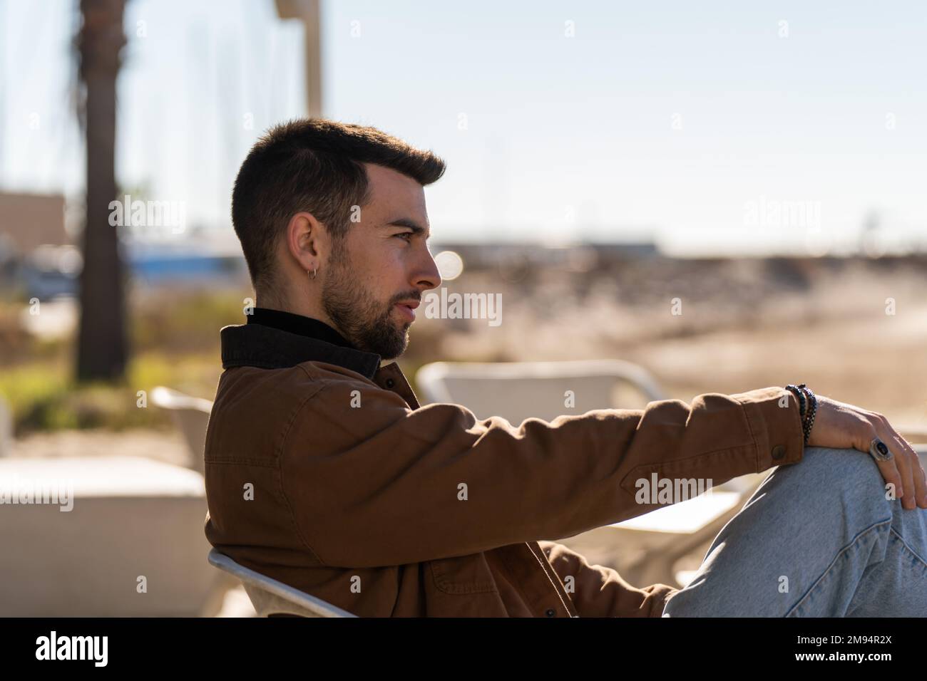 Side view of serious male in outerwear sitting on chair against blurred background of street in sunny weather Stock Photo