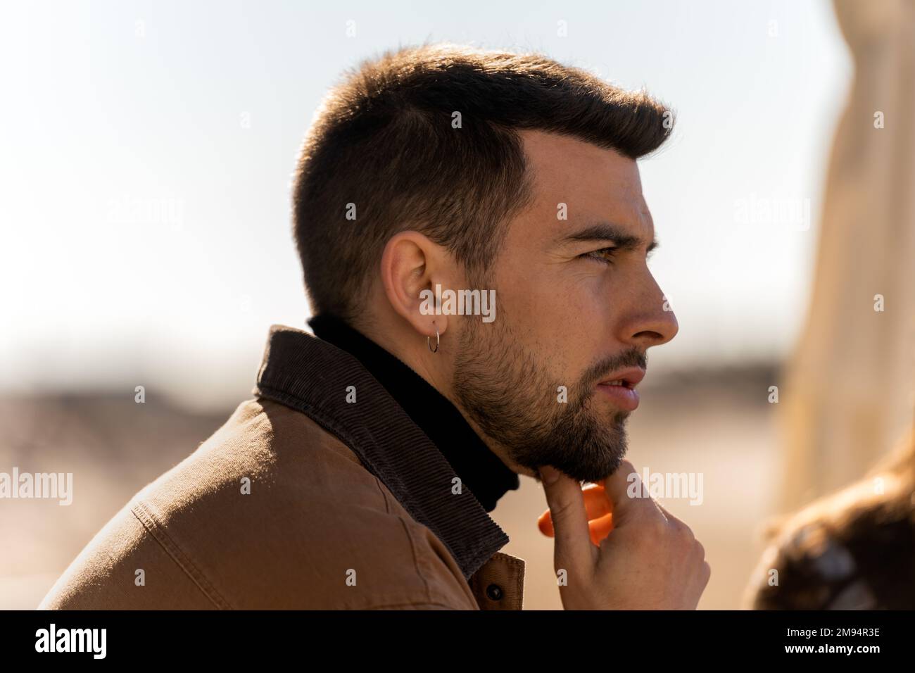 Side view of pensive bearded guy in outerwear with earring looking away thoughtfully while touching chin on blurred background Stock Photo