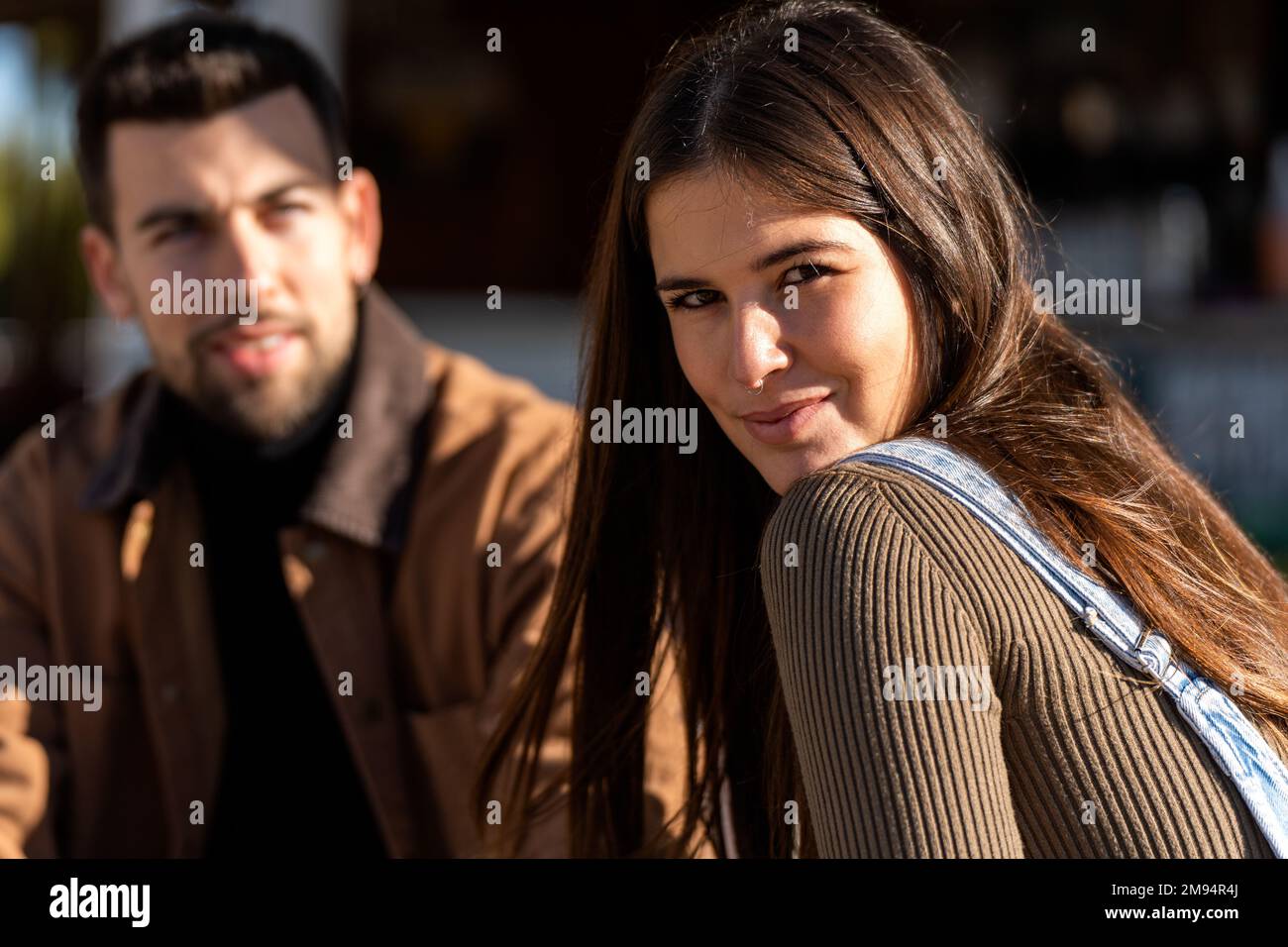 Positive young female in casual clothes with long dark hair looking at camera while sitting with male friend in outerwear on street Stock Photo