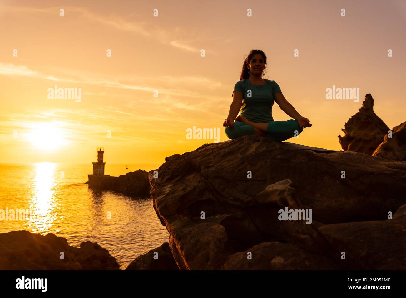 A woman doing meditation and yoga exercises at sunset next to a lighthouse in the sea, healthy and naturist life, outdoor pilates, ardha padmasana Stock Photo