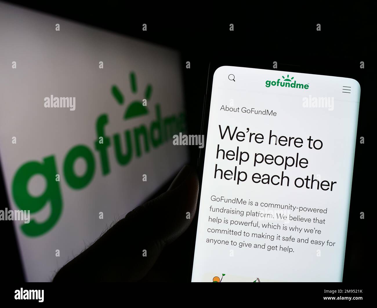 Person holding cellphone with webpage of crowdfunding platform company GoFundMe on screen in front of logo. Focus on center of phone display. Stock Photo