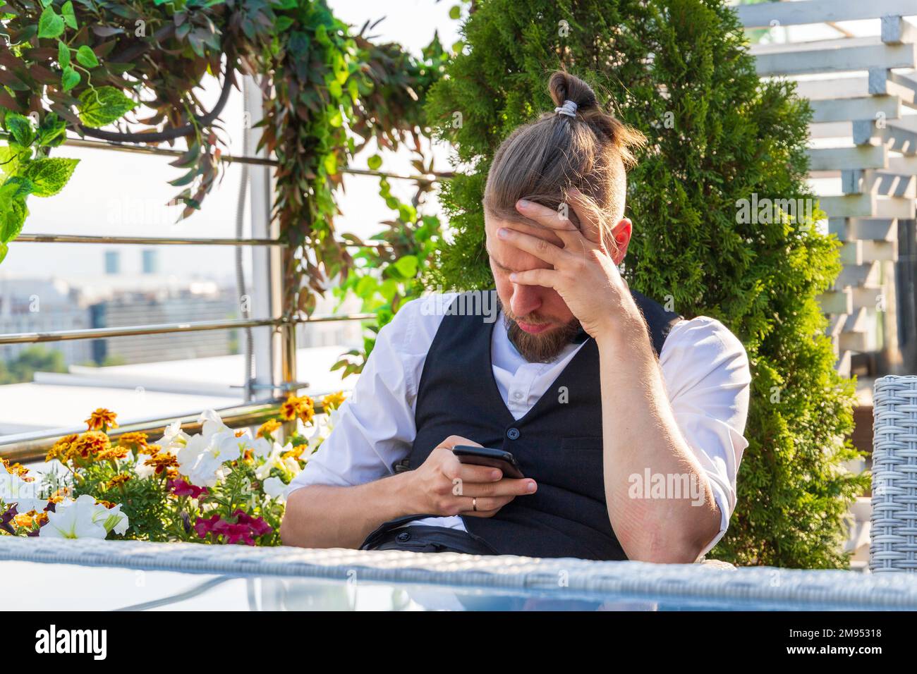 Upset man looking at smartphone holding his head while sitting in penthouse got bad news Stock Photo