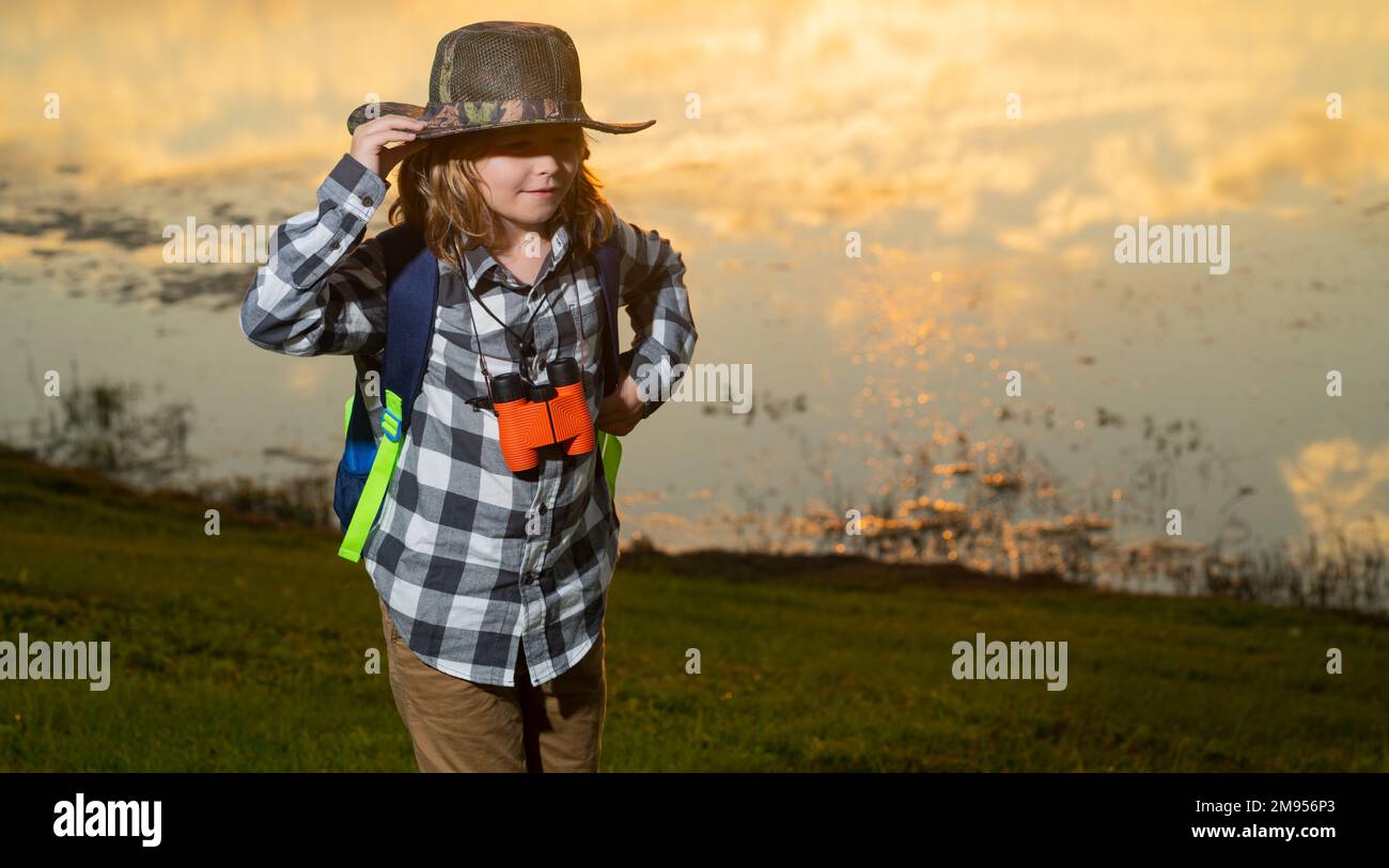 Cute blond kid with binoculars wearing explorer hat and backpack on nature. Child explorer hiking and adventure. Stock Photo